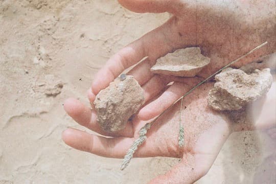 two hands holding small stones and blades of grass over pale sand