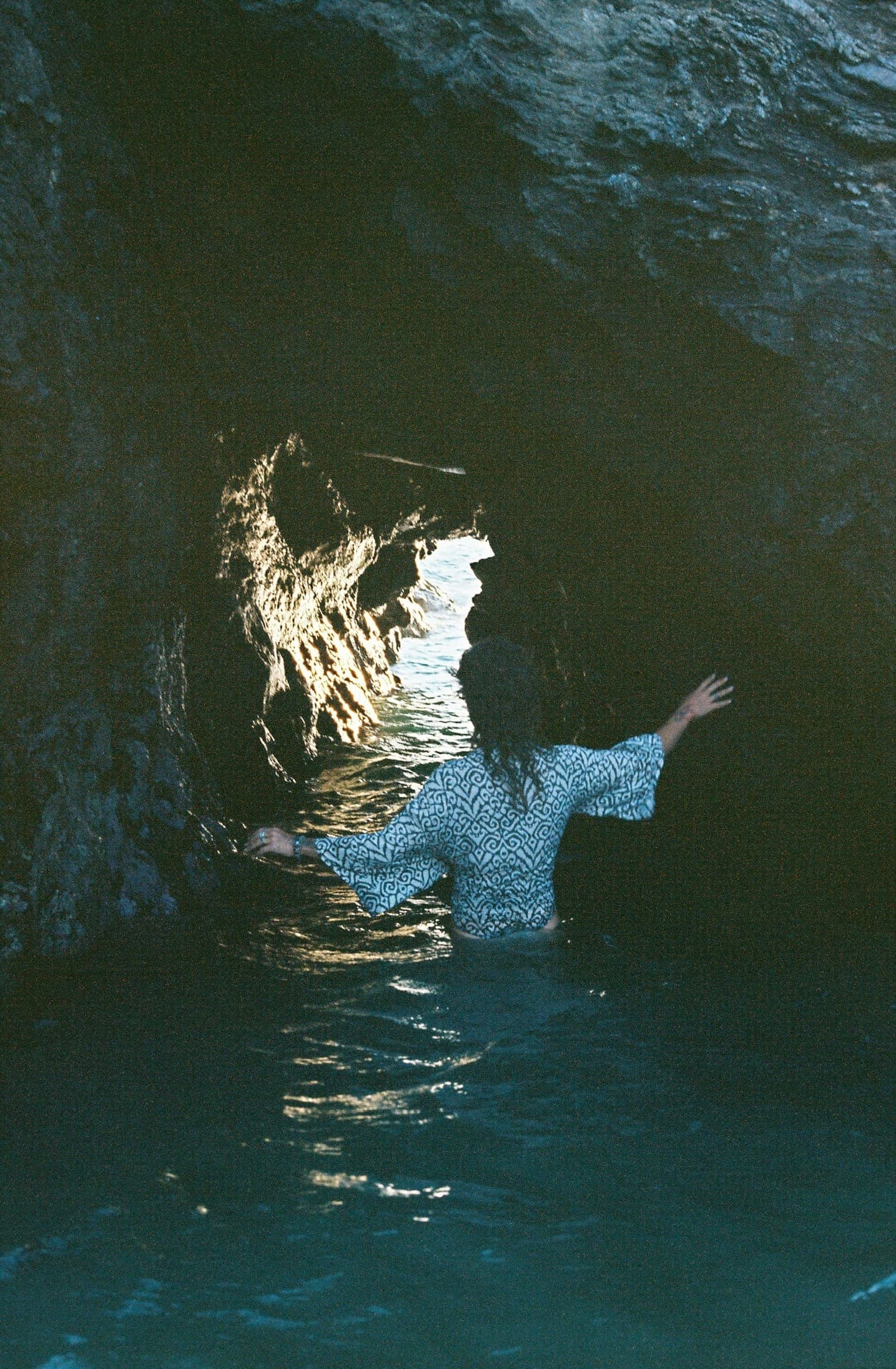 a woman wading into a sea cave, toward the light on the water